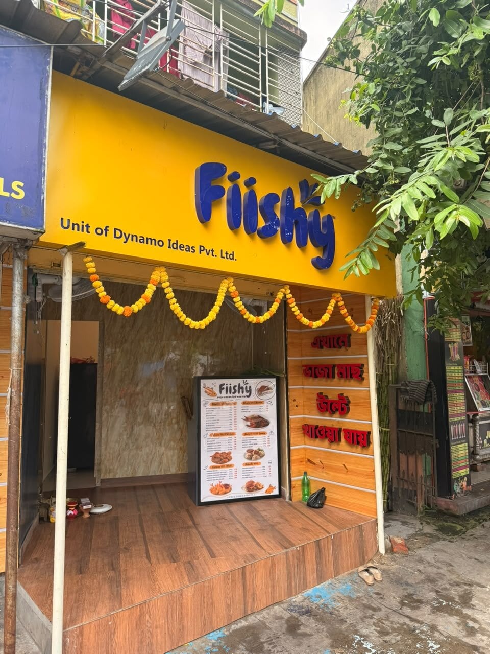 The Kalikapur shopfront by day, yellow board with the blue Fiishy wordmark and marigold garlands over the counter
